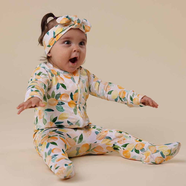 Baby wearing a Sunny Citrus Organic Snuggle Sleepsuit Zip Footie and headband on a beige background