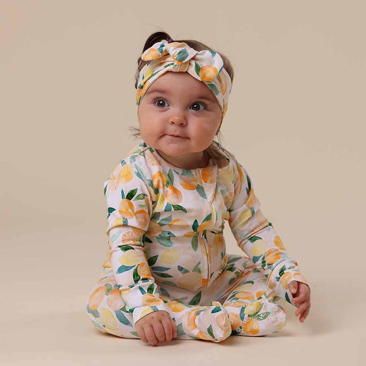 Baby wearing a Sunny Citrus Organic Snuggle Sleepsuit Zip Footie and headband on a beige background