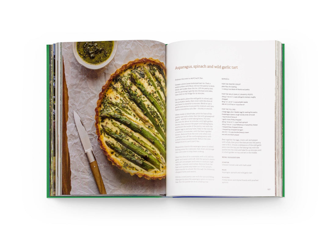 Open cookbook showing a recipe for a green vegetable tart with a knife and small bowl of pesto on a white background.