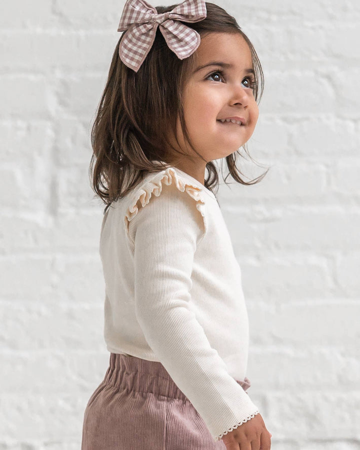 Young girl wearing a white long-sleeve shirt with ruffles and pink checkered bow against a light background
