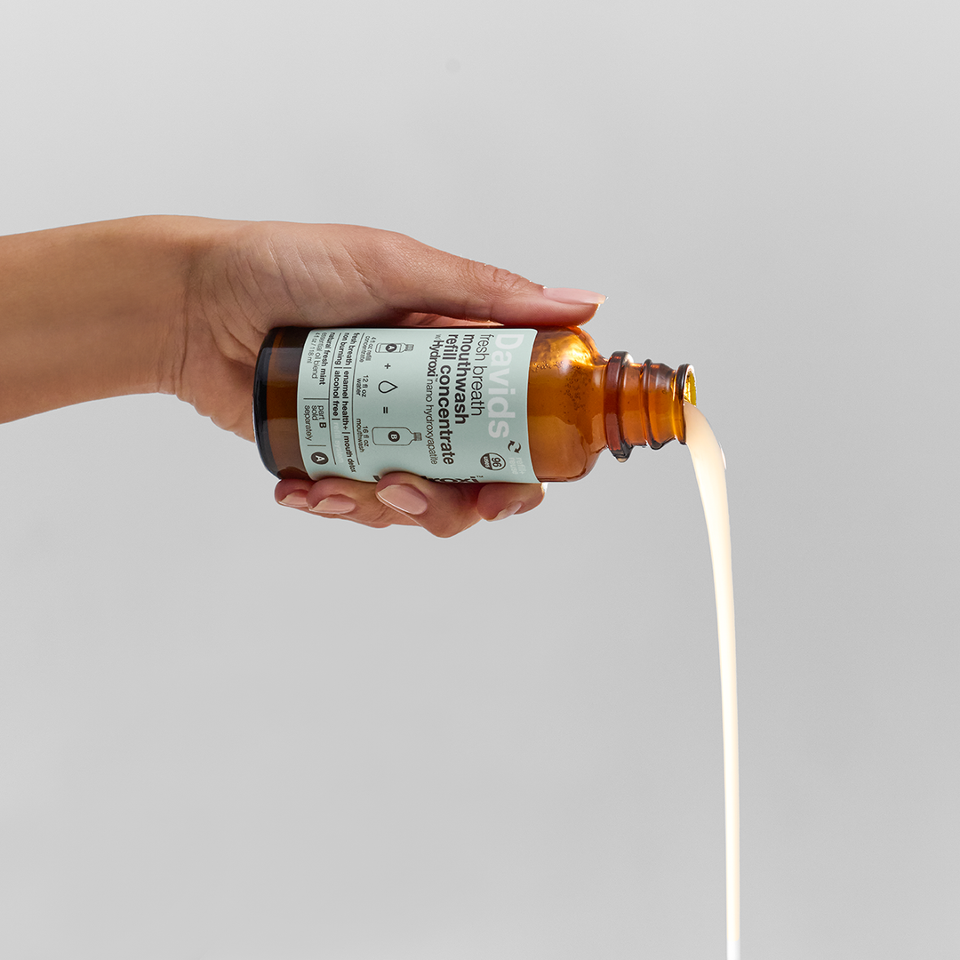 Hand holding Davids mouthwash bottle with a white label, pouring mouthwash onto a light gray background.