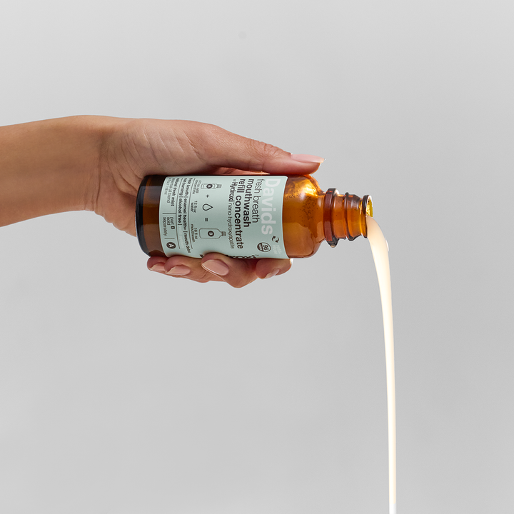 Hand holding Davids mouthwash bottle with a white label, pouring mouthwash onto a light gray background.