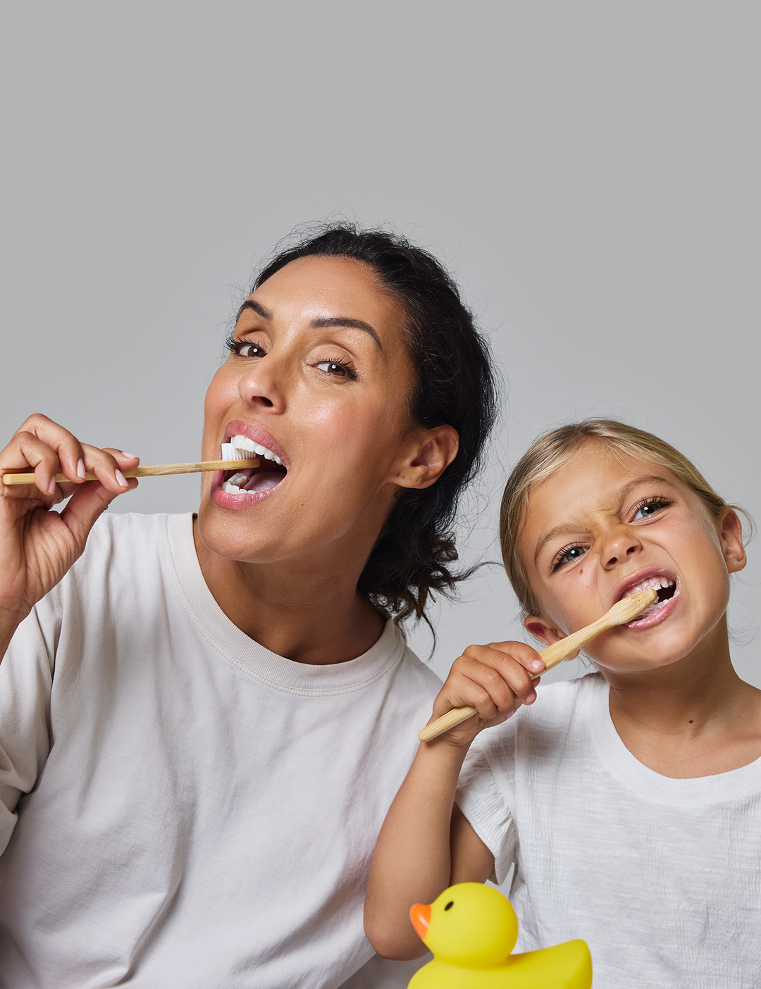 Woman and child brushing their teeth together with a rubber duck on a gray background