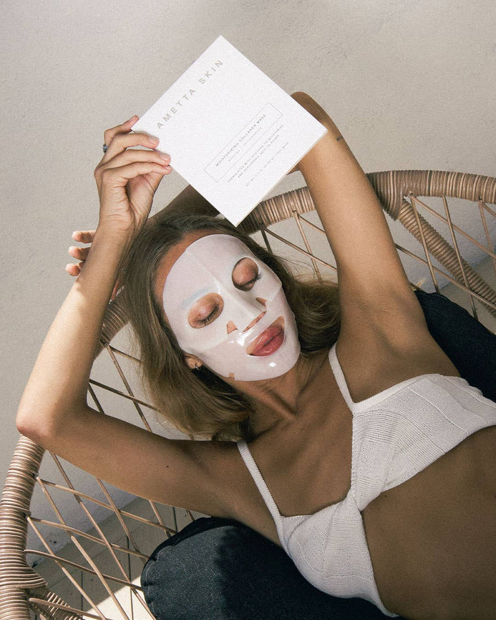 Woman wearing a white face mask and holding a Ametta face mask packaging, sitting on a wicker chair.