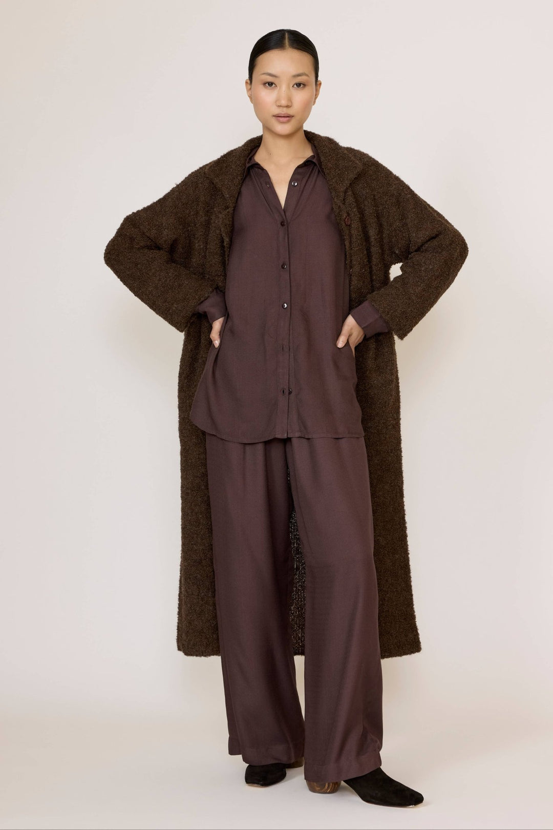 Woman wearing a Laude Everything Shirt in Chocolate Brown with brown pants and a long brown coat with a neutral background