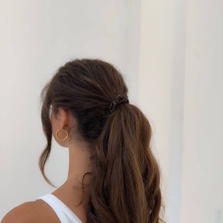 Person with a high ponytail tied back with a brown silk scrunchie wearing gold hoop earrings against a plain background