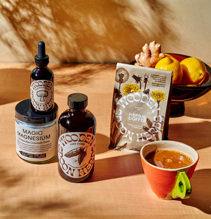 A styled product arrangement on a wooden surface with warm sunlight and shadows. Visible items include a jar labeled Magic Magnesium, a dropper bottle, a glass bottle of Fire Cider and, a bag labeled Wooden on Herbs Herbal Coffee, and a cup of coffee with a green accent handle.