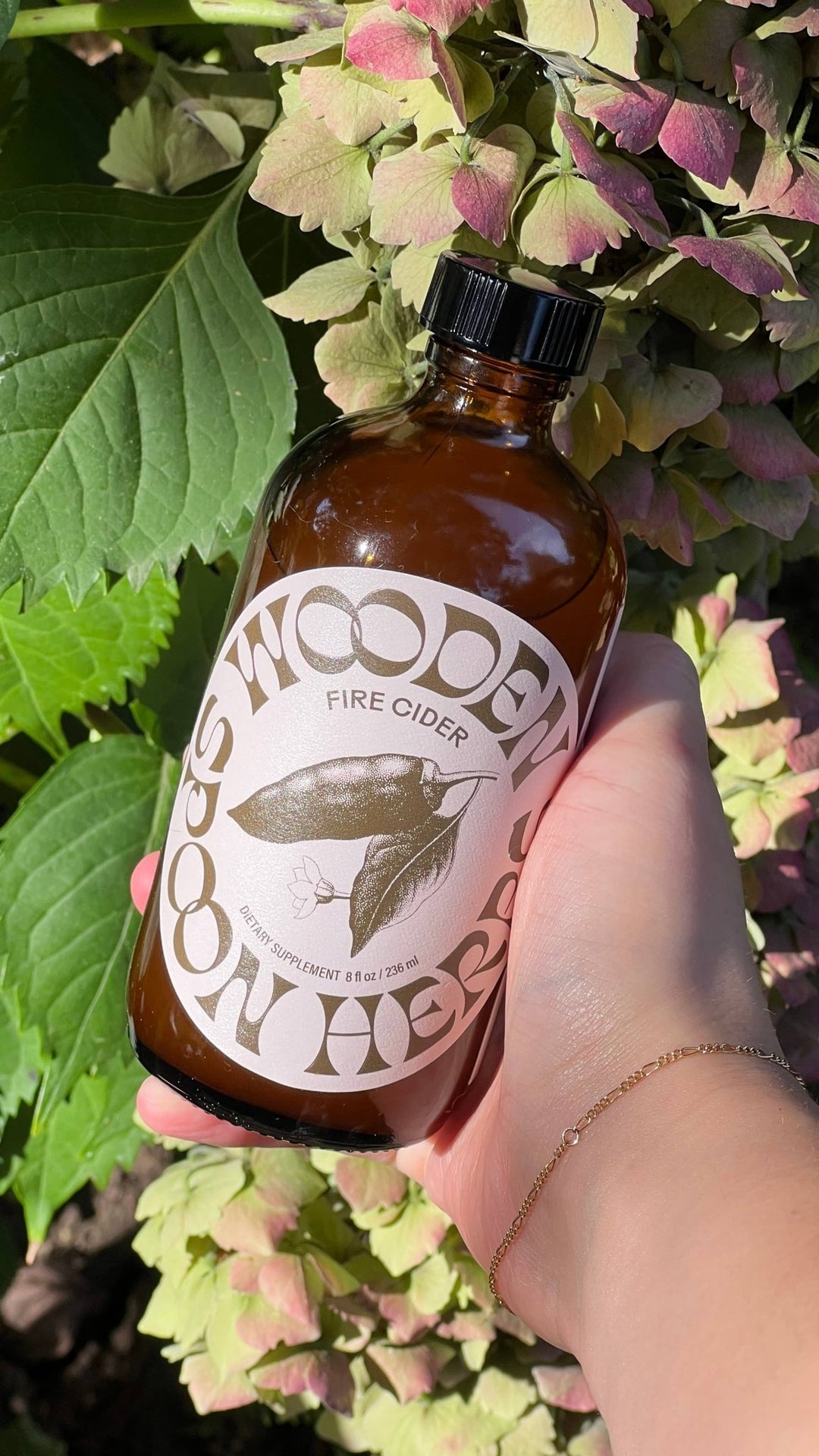 A hand holding an amber glass bottle of Wooden Herbs Fire Cider outdoors, surrounded by green and pink hydrangea flowers. 