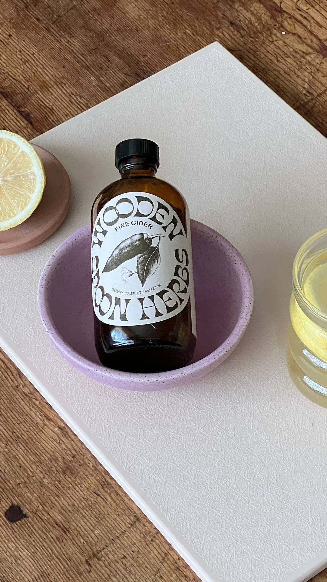 A bottle of Wooden Spoon Fire Cider with a black cap and white label, presented in a light purple ceramic bowl on a white tray over a wooden table.