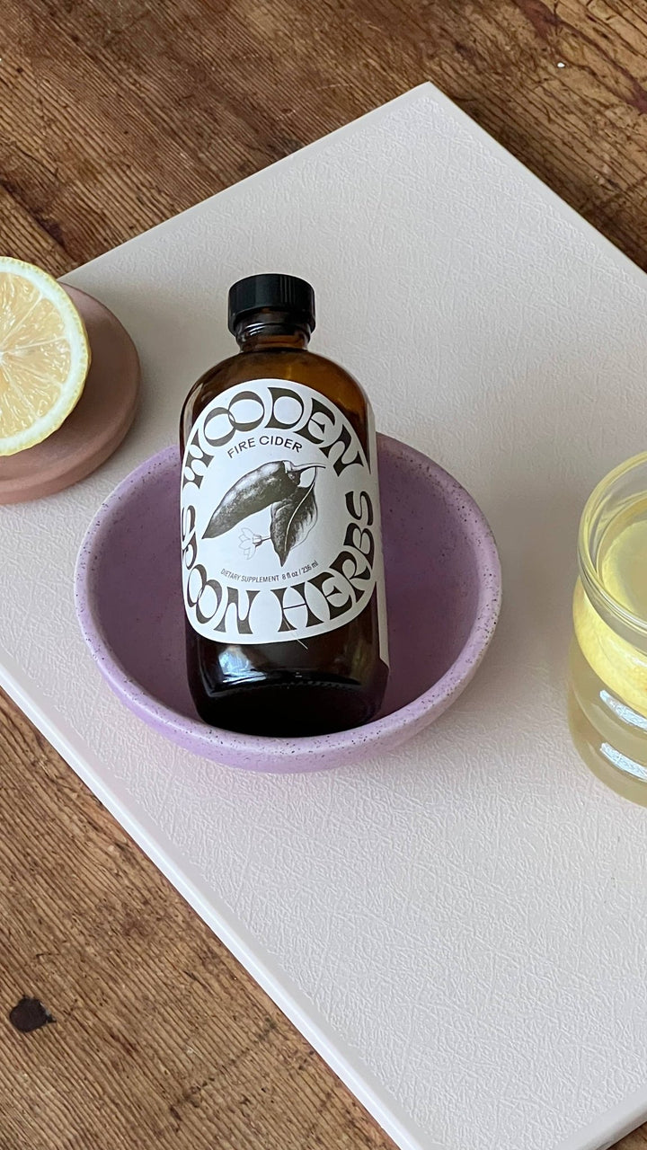A bottle of Wooden Spoon Fire Cider with a black cap and white label, presented in a light purple ceramic bowl on a white tray over a wooden table.