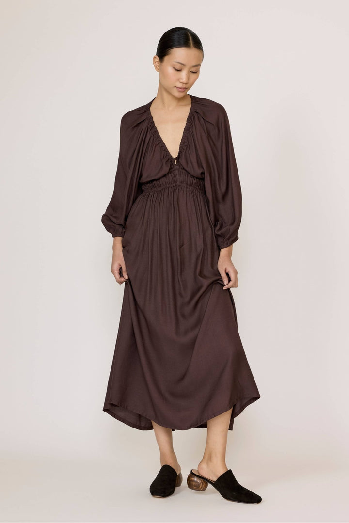 A woman modeling a dark brown midi dress with a deep V-neckline, gathered waist, and long balloon sleeves, paired with black mule shoes against a plain light background.