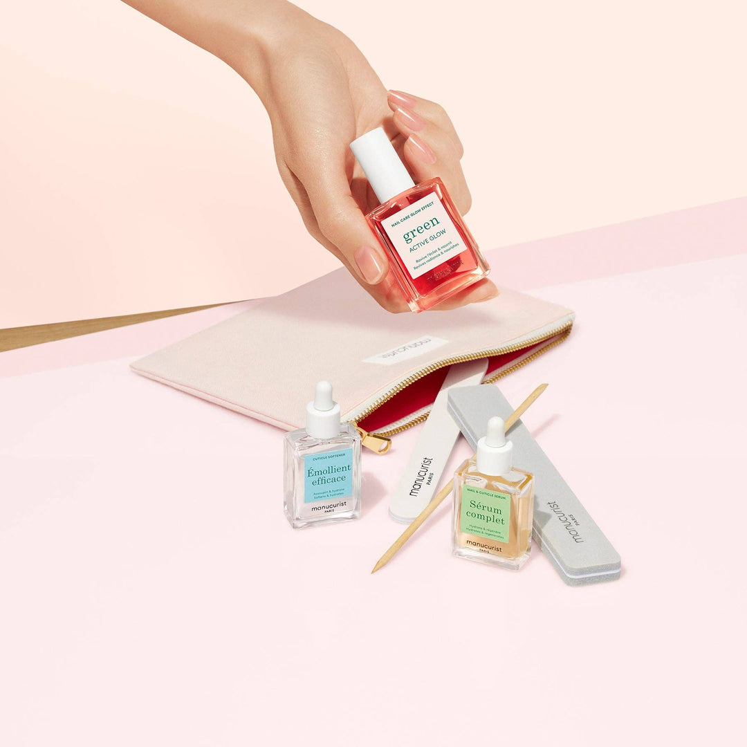 Hand holding a small bottle of nail polish with other nail care bottles and a pouch on a pink background