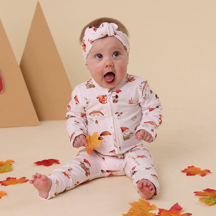 Baby in a Gnomes Organic Snugglesuit Convertible Romper, sitting on a beige surface with colorful leaves.