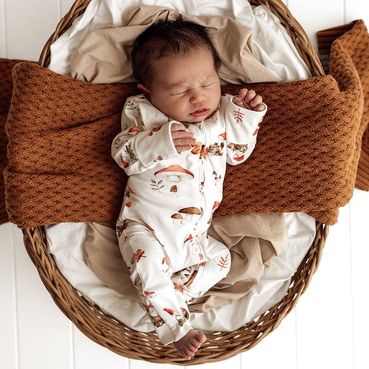 Newborn baby in a wicker basket with brown and beige blankets, wearing a Gnomes Organic Snugglesuit Convertible Romper