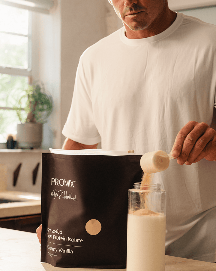 A man in a white t-shirt is shown in a kitchen setting, pouring protein powder from a scoop into a glass bottle. In the foreground, a brown bag of PROMIX Whey Isolate in Creamy Vanilla flavor is clearly visible.