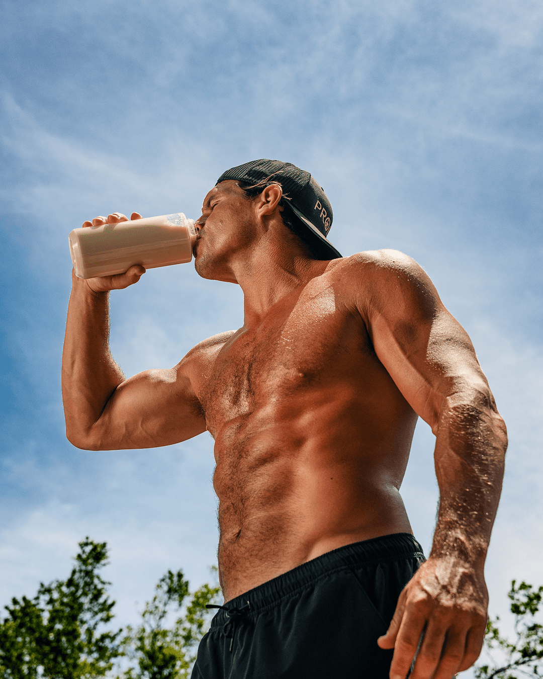 A shirtless muscular man wearing a black cap and black shorts drinks protein from a clear bottle outdoors against a blue sky.