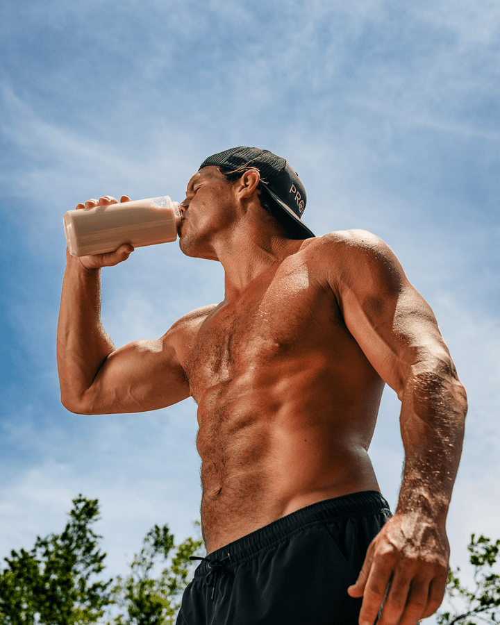 A shirtless muscular man wearing a black cap and black shorts drinks protein from a clear bottle outdoors against a blue sky.