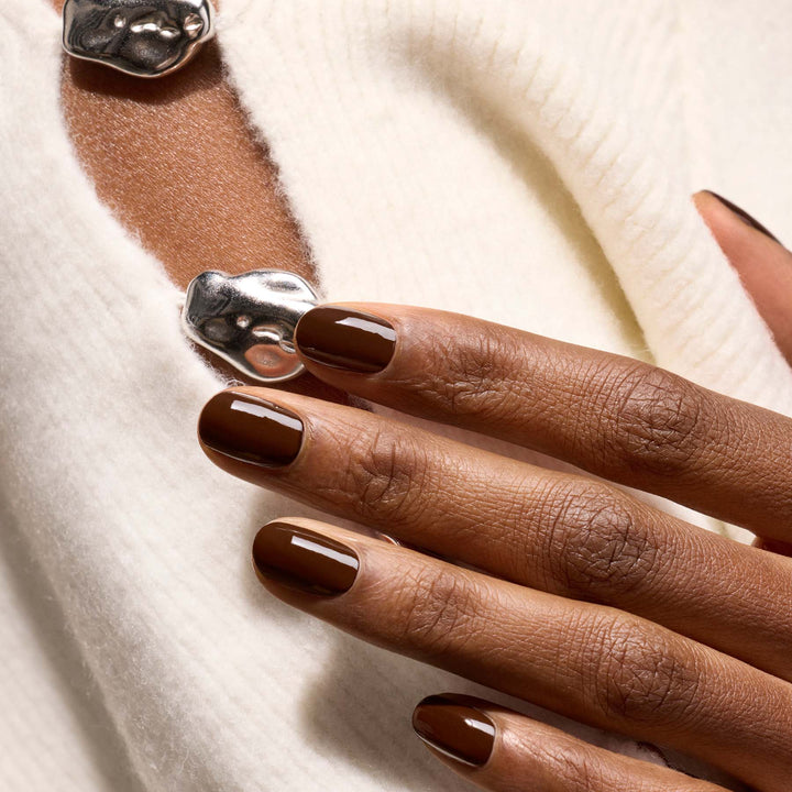 A close-up of a hand with glossy dark brown manicured nails resting on a soft white knit sweater, wearing a chunky silver textured ring.