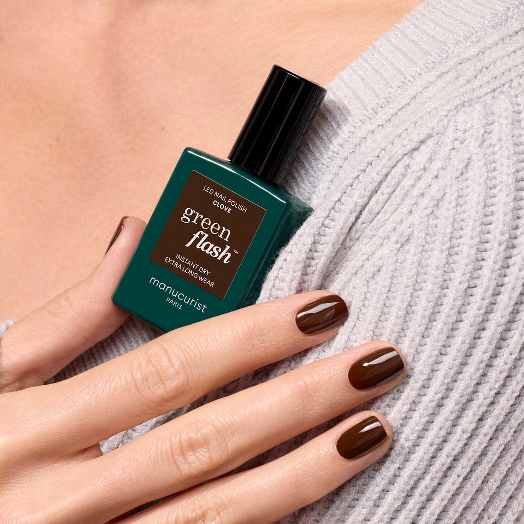 A manucurist bottle of clove LED nail polish held by a hand with glossy dark brown nail polish, resting against a light gray ribbed sweater.