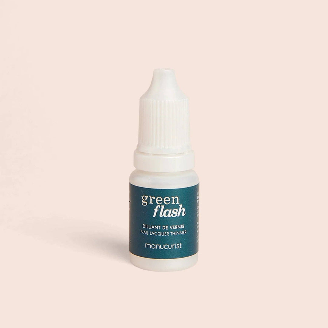A small white bottle of Manucurist Green Flash nail polish thinner with a dark teal label, centered on a solid pale peach background.