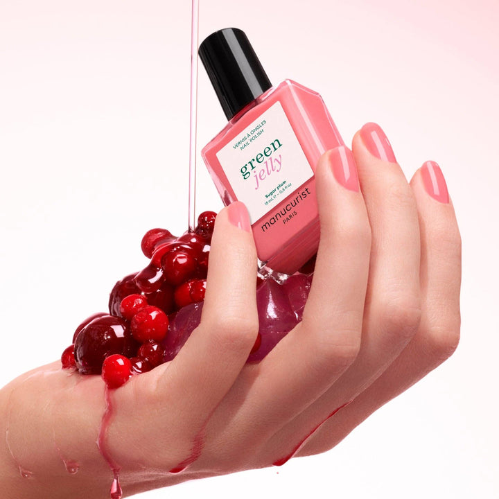 Pink nail polish bottle with red berries and a hand with pink nails on a light pink background