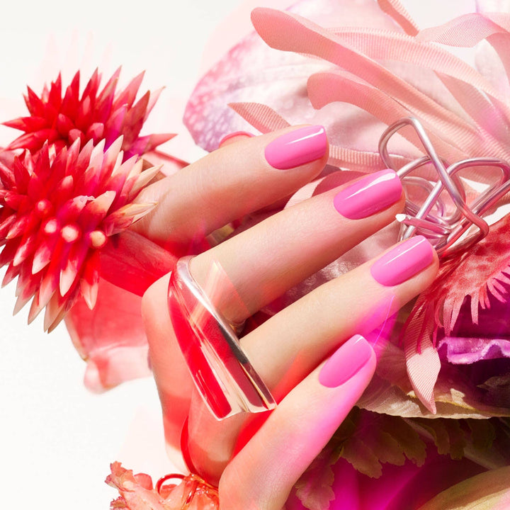 Hand with pink nails holding a bottle of pink nail polish against a floral background