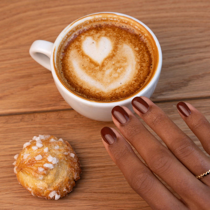 Cappuccino with heart foam and a pastry on a wooden table, with a hand wearing chestnut nail polish reaching towards the pastry.