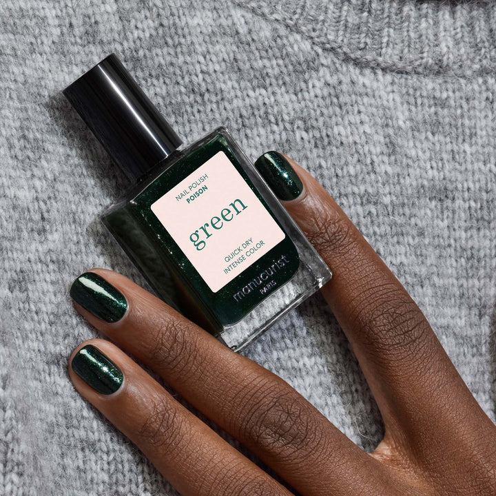 A hand with dark green nail polish holds a bottle of Manucurist poison nail polish against a gray knit background.
