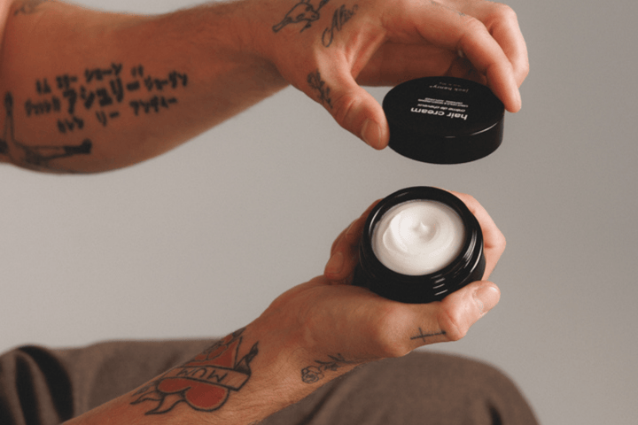Person holding two black containers with hair cream, one open showing the cream.