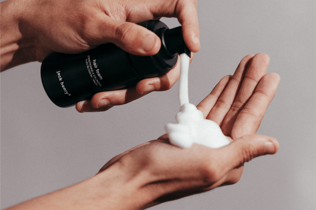 Hand holding a black bottle dispensing hair foam onto another hand against a neutral background