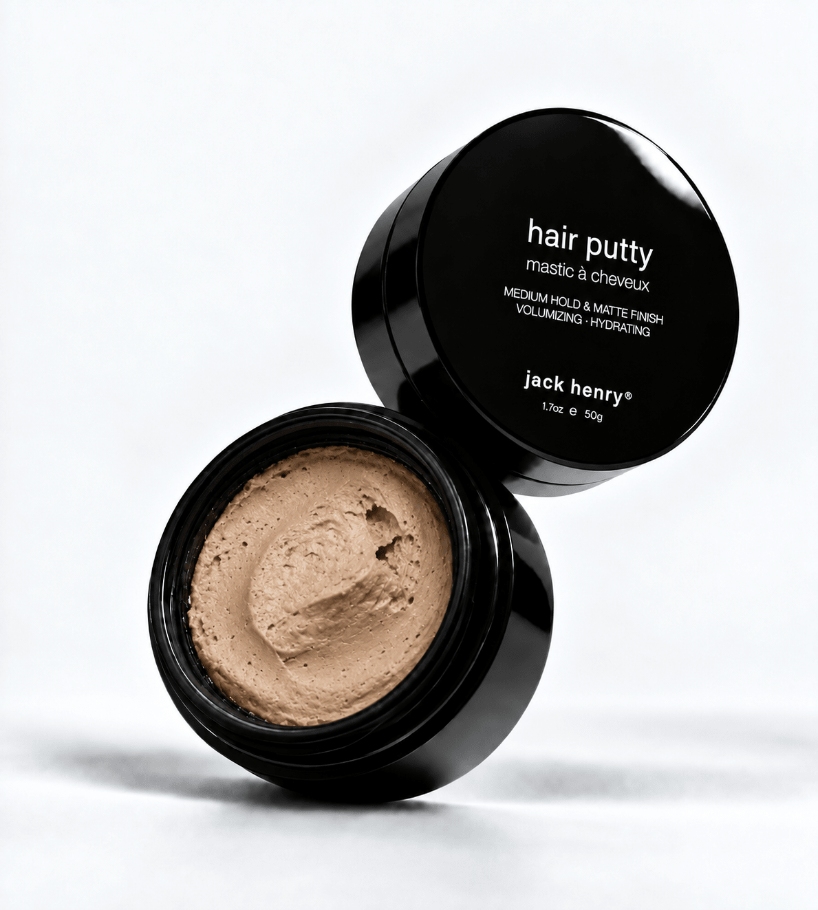 An open black jar of Jack Henry Hair Putty with its lid propped behind it, showing the light tan putty inside. The black lid features white text reading 'hair putty' and 'jack henry'.