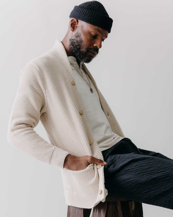 Man wearing a beige cardigan and black beanie sitting on a stool against a plain background