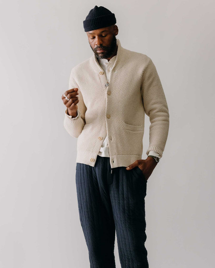 Man wearing a beige cardigan, white shirt, and dark pants against a plain background