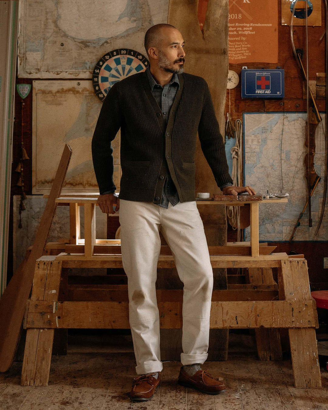 Man standing in a workshop with tools and maps on the wall waering green sweater