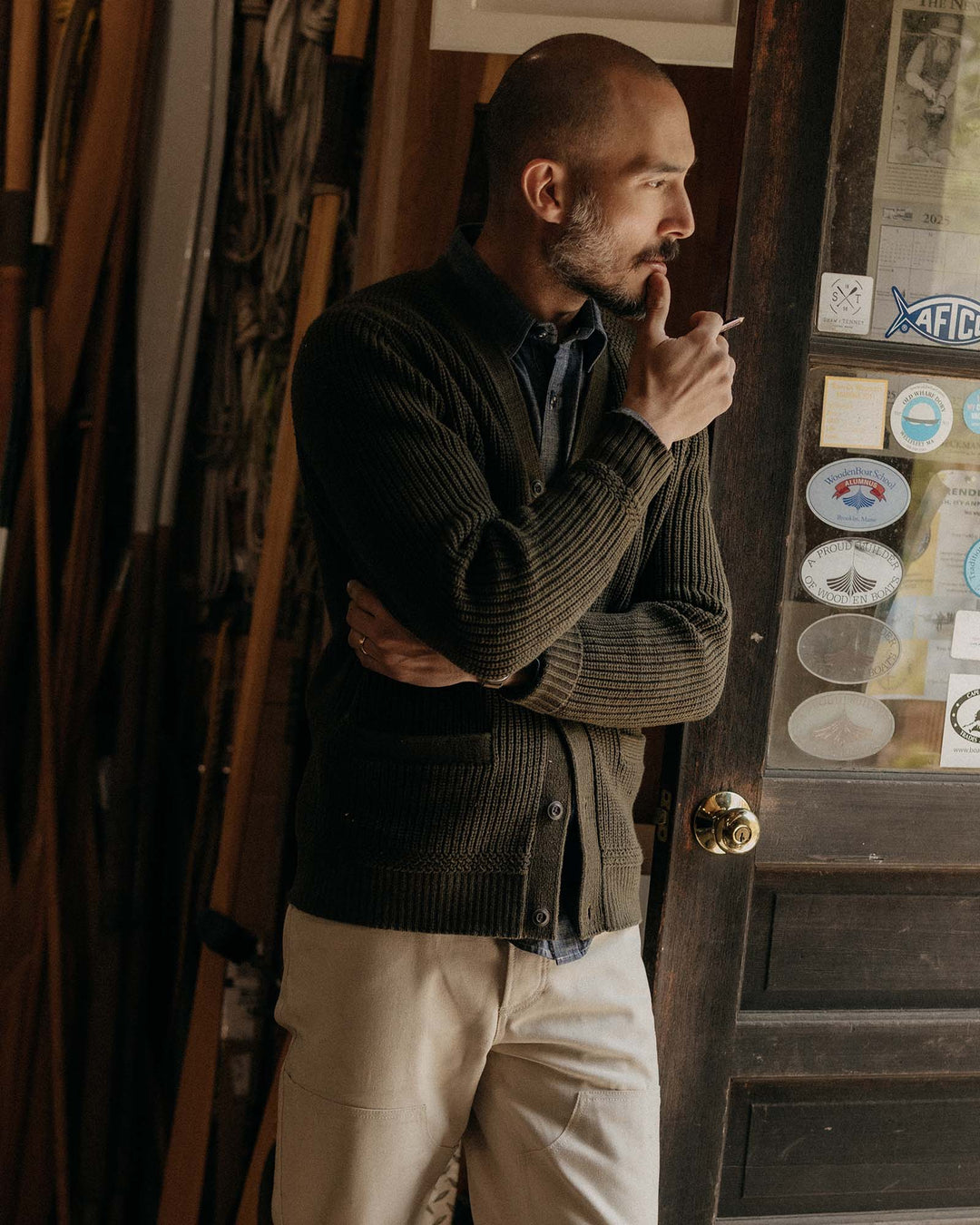 Man in a green sweater and beige pants standing in a room with wooden walls and a door.