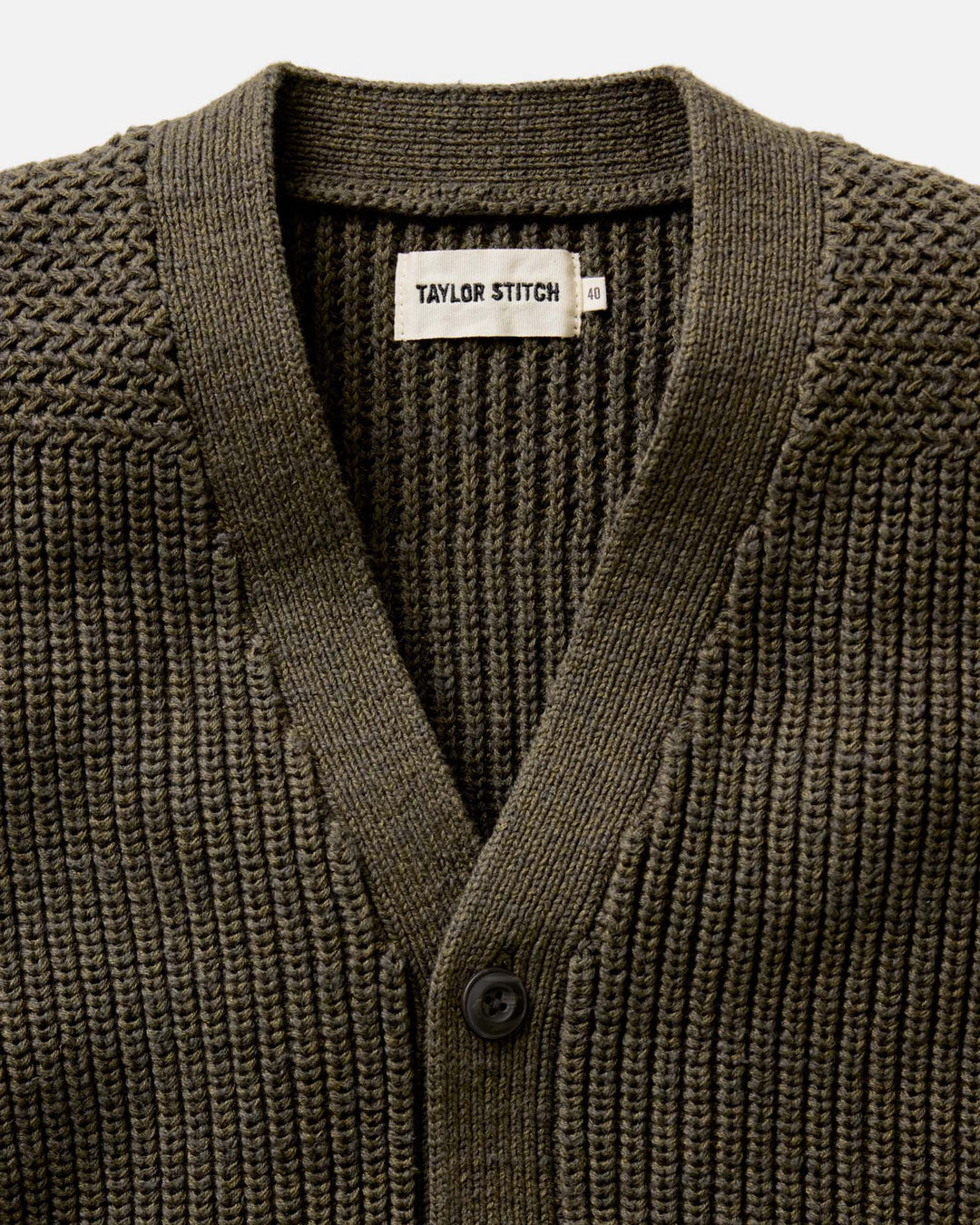 Close-up of a green knitted cardigan with 'Taylor Stitch' label on a light gray background