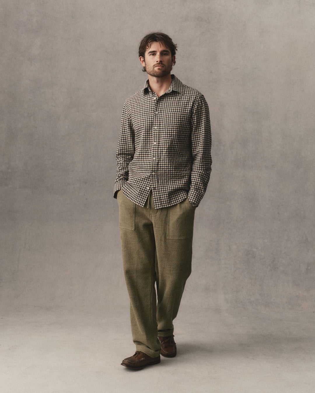 A man stands against a textured gray studio backdrop wearing The California Shirt in Breaker Gingham, olive green trousers, and brown leather shoes.