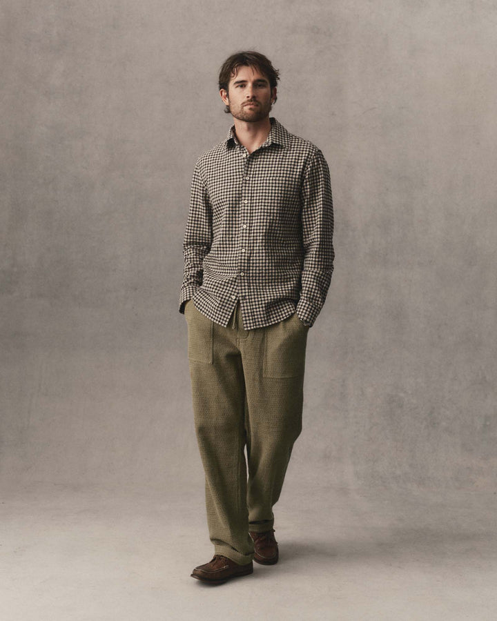 A man stands against a textured gray studio backdrop wearing The California Shirt in Breaker Gingham, olive green trousers, and brown leather shoes.