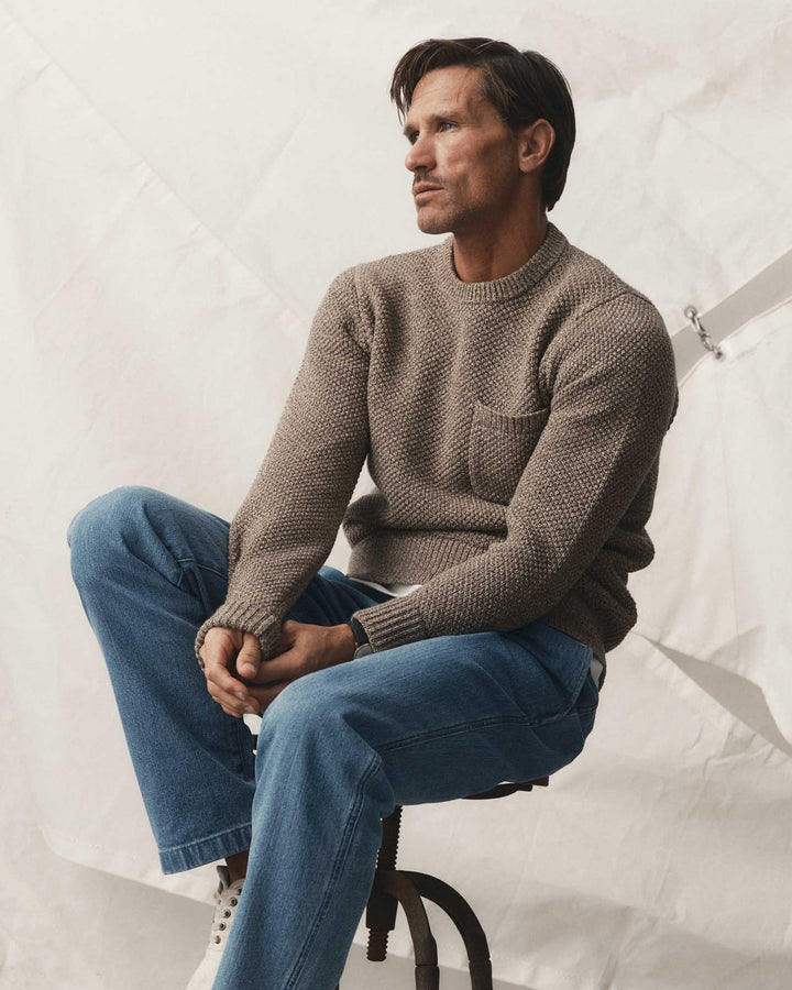 A man wearing the Crawford Crew Sweater in Coffee Twist and blue jeans sits on a stool against a white studio backdrop.