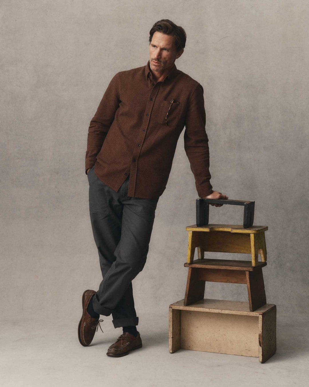 A man in The Jack Shirt in Timber Twist Waffle and dark trousers leans against a small wooden step stool with a black handle in a studio setting with a textured gray background.