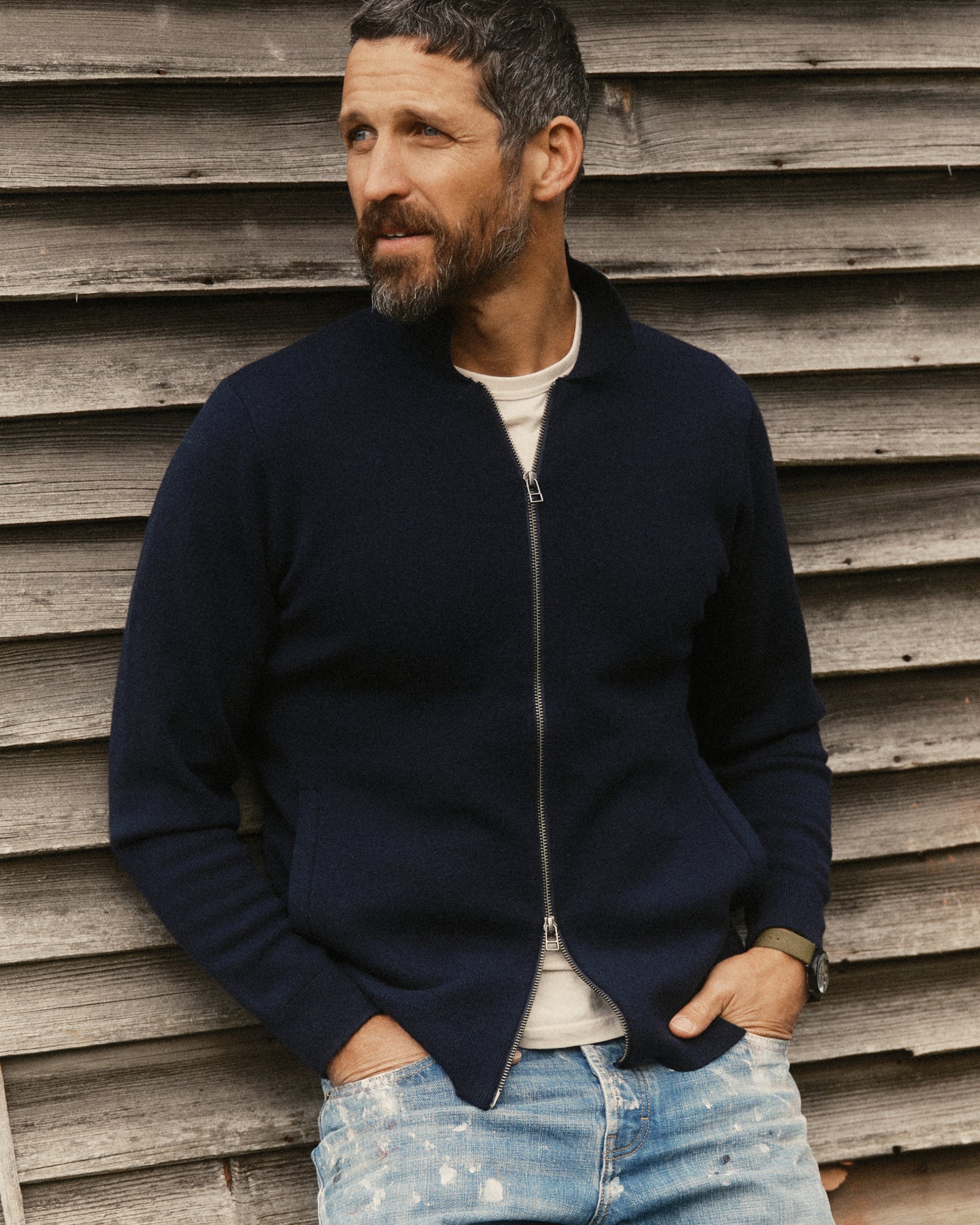 A man wearing Taylor Stitch's Portola Sweater Jacket in Midnight Merino over a white T-shirt and light blue jeans, standing against a rustic wooden plank wall.
