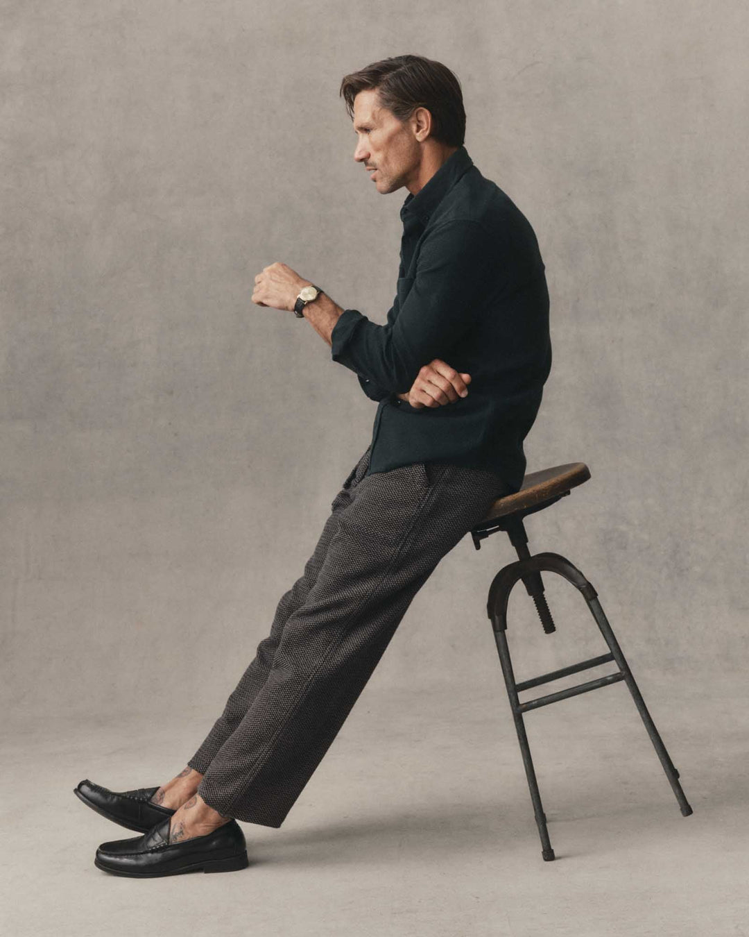 A man in a Jack Shirt in Forest Waffle and gray trousers sits on a black metal stool, looking down at his watch. The background is a textured gray studio wall.