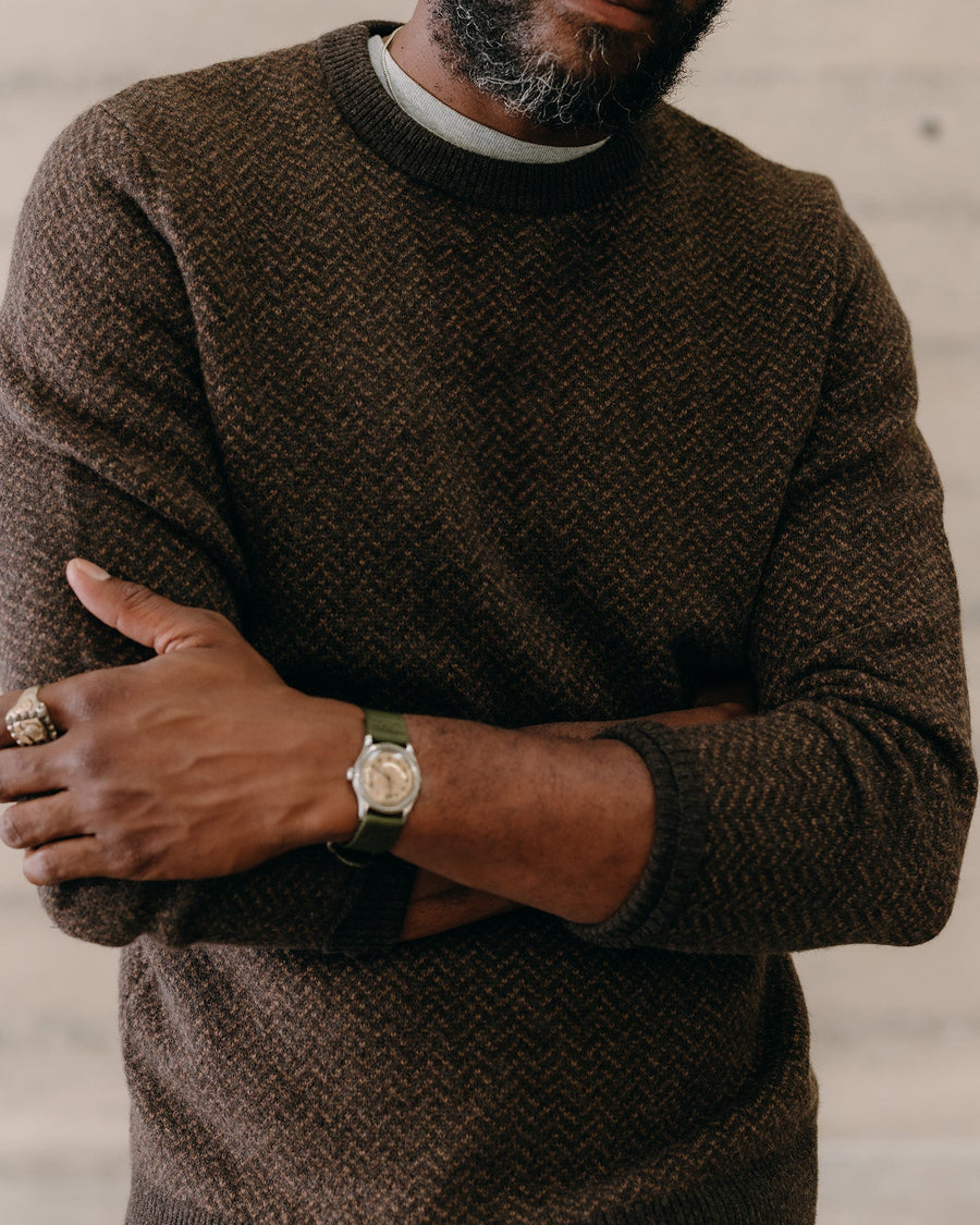 Man wearing a brown sweater with a watch on his wrist, standing against a neutral background