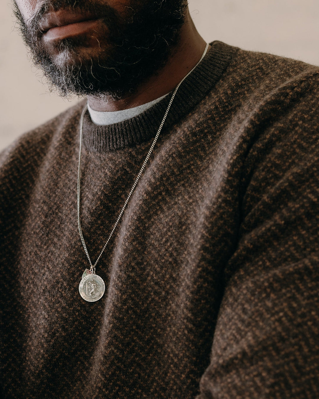 Man wearing a brown sweater with a necklace featuring a circular pendant.