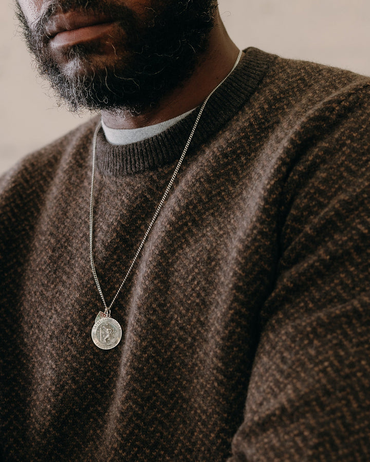 Man wearing a brown sweater with a necklace featuring a circular pendant.