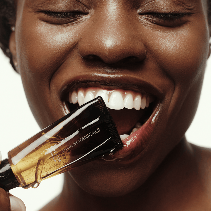 Close-up of a woman with a bottle of True Botanicals lipid lip oil by her mouth.