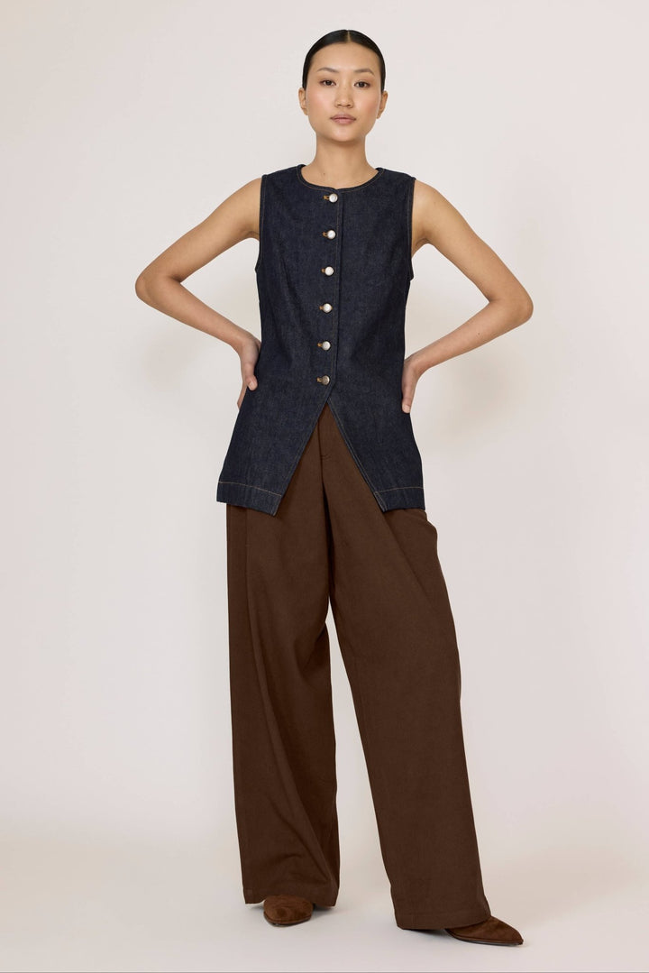 Woman wearing a Laude Longline Vest in Dark Indigo with brown pants over a plain background