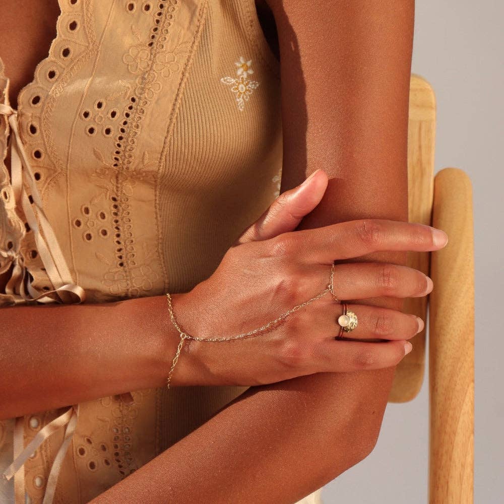 Close-up of a person wearing a gold ring and hand chain, sitting on a wooden chair.