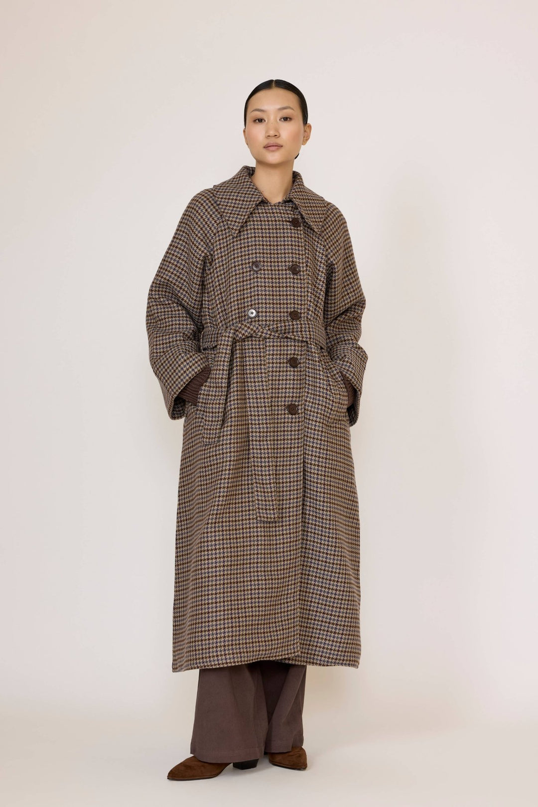 Person wearing a long brown checkered coat and brown pants underneath on a white background