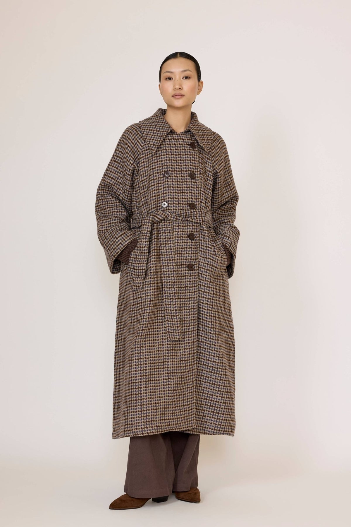 Person wearing a long brown checkered coat and brown pants underneath on a white background